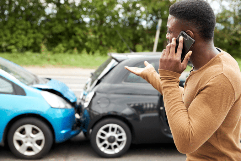 A man holding a phone to his left cheek, appearing to make a call, while gesturing with his right hand to demonstrate a collision between two cars.