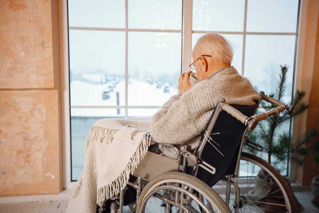 Old man sitting in a a wheelchair near big window looking out to the snow and drinking a cup of tea