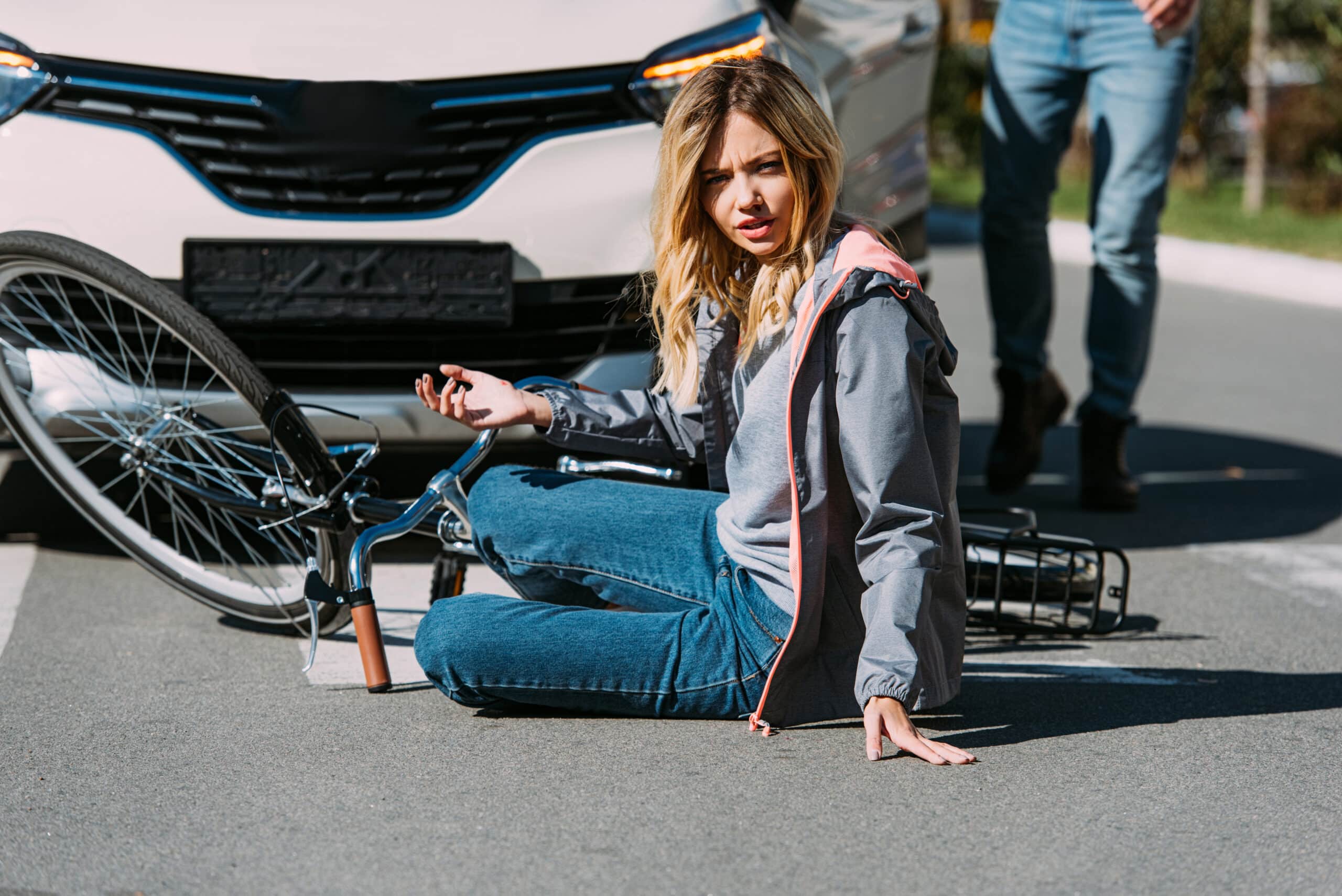 partial view of injured woman on road after bicycle and car accident with car driver behind