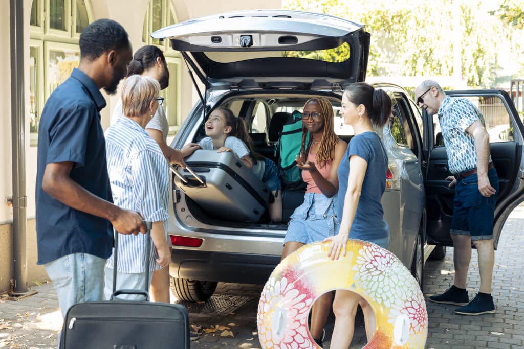 Small kid travelling on vacation with family, preparing to go to seaside holiday during summer. Diverse people and friends loading suitcase and trolley in car trunk, leaving on journey trip.